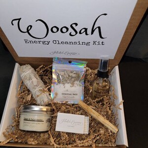 May include: A brown cardboard box with the words "Woosah Energy Cleansing Kit" printed on the lid. The box contains a white sage smudge stick, a small bag of sage and lavender for a spiritual bath, a bottle of energy cleansing spray, a palo santo stick, and a box of matches. The box is filled with brown shredded paper. The words "Regular Kit" are printed on the bottom of the box.