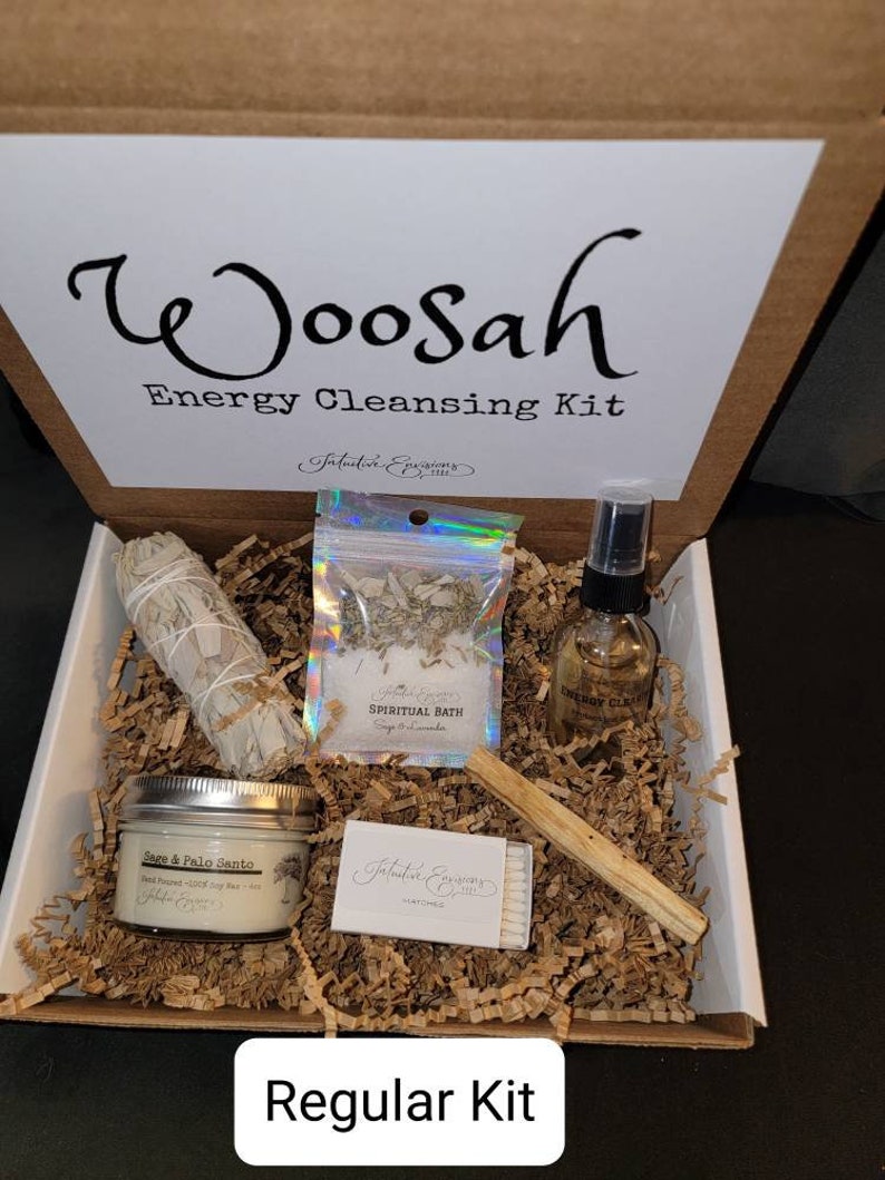 May include: A brown cardboard box with the words "Woosah Energy Cleansing Kit" printed on the lid. The box contains a white sage smudge stick, a small bag of sage and lavender for a spiritual bath, a bottle of energy cleansing spray, a palo santo stick, and a box of matches. The box is filled with brown shredded paper. The words "Regular Kit" are printed on the bottom of the box.