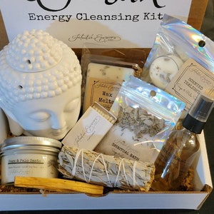 May include: A white box filled with items for an energy cleansing kit. The box has the words "Woosah Energy Cleansing Kit" printed on the top. The kit includes a white ceramic Buddha head, a white candle, a bag of white sage, a bag of palo santo, a bag of bath salts, a bottle of essential oil, a pack of matches, and a wax melt.