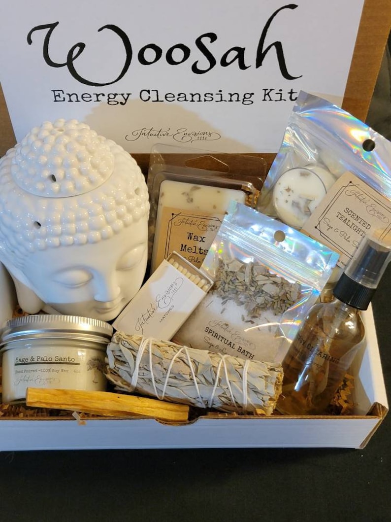 May include: A white box filled with items for an energy cleansing kit. The box has the words "Woosah Energy Cleansing Kit" printed on the top. The kit includes a white ceramic Buddha head, a white candle, a bag of white sage, a bag of palo santo, a bag of bath salts, a bottle of essential oil, a pack of matches, and a wax melt.