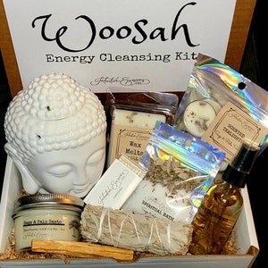 May include: A white box with the words "Woosah Energy Cleansing Kit" on the front. The box contains a white Buddha head incense burner, a white wax melt, a white tea light, a bag of sage, a bundle of white sage smudge sticks, a bottle of essential oil, and a stick of palo santo wood. The box also has the words "Deluxe Kit" on the bottom.