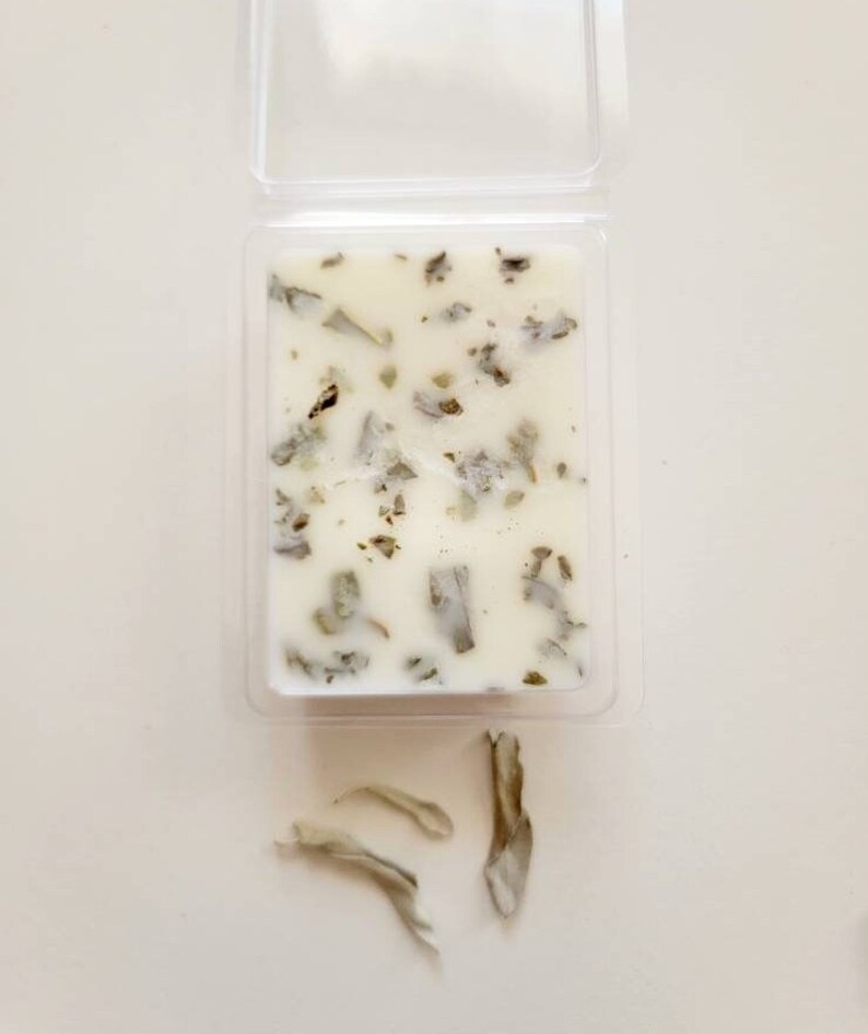 Pu&ograve; includere: Un fondente di cera bianco con foglie di salvia secche incorporate. Il fondente di cera &egrave; in un contenitore di plastica trasparente. Ci sono tre pezzi di foglie di salvia secche su una superficie bianca sotto il contenitore.