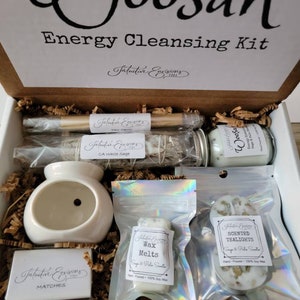May include: A starter kit for energy cleansing with the words "Woosah Energy Cleansing Kit" on the box. The kit includes a white ceramic oil burner, a small box of matches, a bag of wax melts, a bag of scented tealights, a small jar of white sage, and a wooden smudge stick.