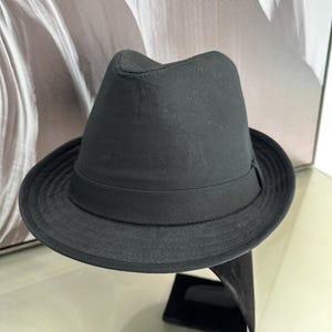 Könnte beinhalten: Ein schwarzer Fedora-Hut mit strukturierter Krempe und einem Ripsband. Der Hut wird auf einem schwarzen Ständer präsentiert, der sein klassisches Design hervorhebt. Die dunkle Farbe und die schlichten Linien des Hutes verleihen ihm eine zeitlose Ausstrahlung.