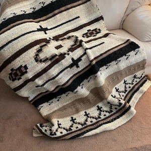 Könnte beinhalten: Eine gehäkelte Wolldecke mit einem geometrischen Muster in Braun-, Schwarz- und Weißtönen. Die Decke ist gefaltet und über ein weißes Sofa gelegt.