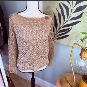 Pode incluir: Um suéter de crochê bege com mangas compridas e decote canoa. O suéter apresenta um design de tecido aberto com pequenos enfeites brilhantes. A peça de roupa é exibida em um manequim.