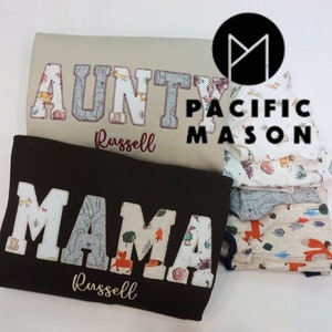 May include: Two brown shirts with the words "AUNTY" and "MAMA" spelled out in white letters with a woodland animal print design. The shirts are personalized with the name "Russell" in maroon embroidery. The shirts are stacked on top of a stack of fabric with a woodland animal print design. The top fabric has the words "PACIFIC MASON" in black text.