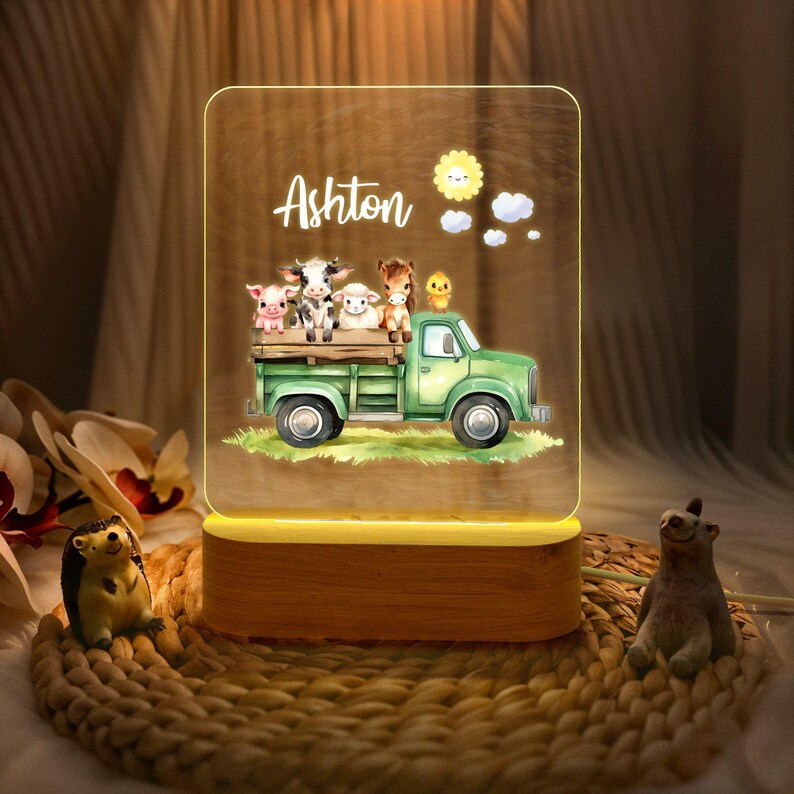 May include: A personalized night light with a wooden base and a clear acrylic panel featuring a green truck carrying farm animals. The truck is carrying a pig, a cow, a sheep, and a chick. The name Ashton is written on the panel.