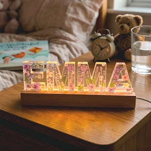 May include: A lighted wooden night light spelling "EMMA" with clear resin letters filled with pink and purple flowers. The night light sits on a wooden surface, with a book, teddy bear, and alarm clock in the background.