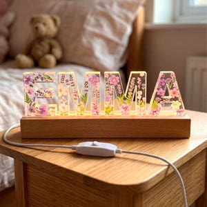 May include: A decorative illuminated sign spelling "EMMA" with clear resin letters filled with pink and purple flowers and greenery. The letters sit on a wooden base with a power cord and switch. The sign is on a wooden nightstand.