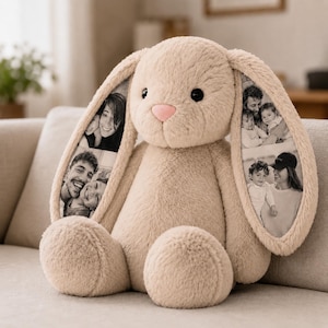 Puede incluir: Un peluche de conejo beige con orejas largas y caídas. La superficie interior de cada oreja presenta fotografías en blanco y negro de personas. El conejo tiene una textura suave y esponjosa, ojos redondos oscuros y una pequeña nariz rosa.