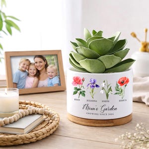 May include: A white ceramic planter with a wooden base, holding a green succulent. The planter is decorated with floral illustrations and the text "Mom's Garden" and names. A framed family photo and a candle are in the background.