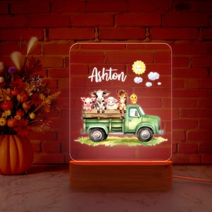 May include: A wooden night light with a clear acrylic panel featuring a green truck with a white bed carrying a cow, pig, lamb, horse, and a chick. The acrylic panel has a white sun with a smiling face and clouds. The text "Ashton" is printed in white on the acrylic panel.