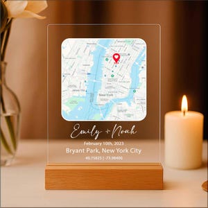 May include: A clear acrylic plaque with a map of New York City, featuring a red marker. The plaque includes the names "Emily + Noah", date, location, and coordinates. It rests on a wooden base, with a lit candle in the background.