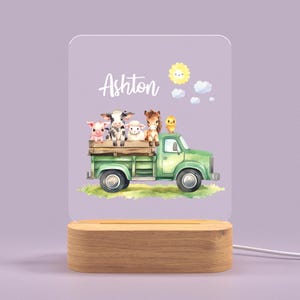 May include: A green toy truck with a white cab and a wooden bed. The truck is carrying a pig, a cow, a lamb, a horse, and a chick. The truck is on a green grassy field with a blue sky and a yellow sun. The name Ashton is written on the truck.