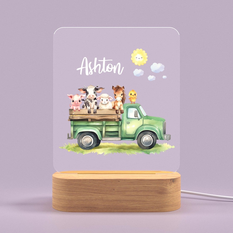 May include: A green toy truck with a white cab and a wooden bed. The truck is carrying a pig, a cow, a lamb, a horse, and a chick. The truck is on a green grassy field with a blue sky and a yellow sun. The name Ashton is written on the truck.