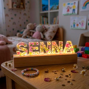 May include: A decorative illuminated sign spelling "SENNA" in clear resin letters filled with colorful flowers. The letters sit on a wooden base, casting a warm glow. A colorful bracelet and scattered flower petals are on the table.