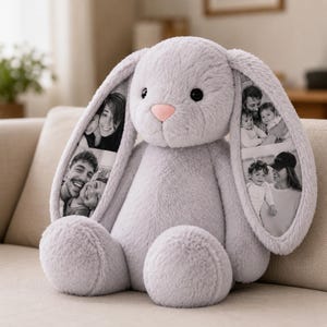 Puede incluir: Un conejo de peluche de color gris claro con orejas largas y caídas. La superficie interior de cada oreja presenta fotografías en blanco y negro de personas. El conejo tiene una textura suave y esponjosa, ojos redondos oscuros y una nariz rosa.