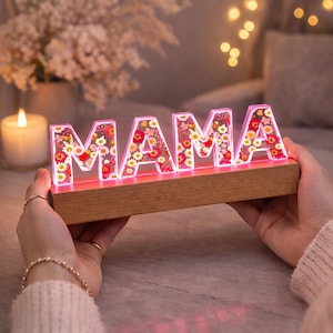 Puede incluir: Un letrero decorativo iluminado con la palabra "MAMA" y detalles florales. Las letras están llenas de flores coloridas y colocadas sobre una base de madera. El letrero se ilumina con un brillo rosado, creando un ambiente cálido.