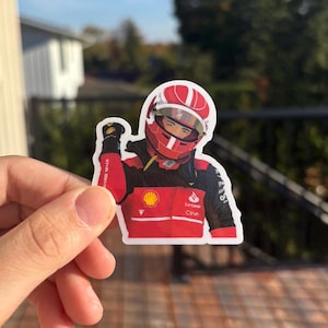 May include: A sticker featuring a Formula 1 driver in a red racing suit and helmet. The sticker has a white border and includes sponsor logos. The driver is celebrating with a raised fist.