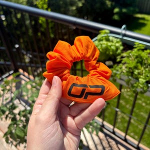 Peut inclure: Chouchou orange vif avec les lettres noires "OP" imprim&eacute;es sur le tissu. Le chouchou est fait d'une mati&egrave;re douce et est rassembl&eacute; autour d'un &eacute;lastique. En arri&egrave;re-plan, on aper&ccedil;oit des plantes vertes et une balustrade noire.