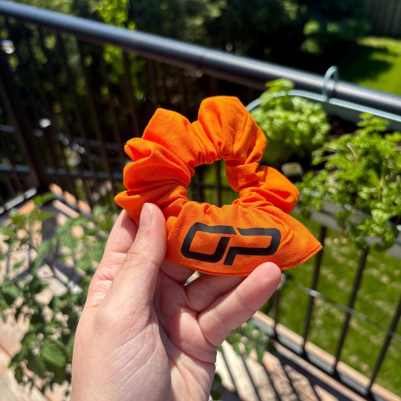 Peut inclure: Chouchou orange vif avec les lettres noires "OP" imprim&eacute;es sur le tissu. Le chouchou est fait d'une mati&egrave;re douce et est rassembl&eacute; autour d'un &eacute;lastique. En arri&egrave;re-plan, on aper&ccedil;oit des plantes vertes et une balustrade noire.