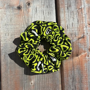 Peut inclure: Un chouchou en tissu avec un motif abstrait répétitif de formes organiques vert citron sur fond noir. Le design comprend un petit logo blanc avec la lettre G imprimée sur la surface du tissu, créant un aspect à fort contraste visuel.