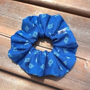 Peut inclure: Un chouchou bleu avec un motif répété du chiffre 55 et une petite étiquette blanche. Le tissu est bleu vif avec une impression bleu clair. Le chouchou est circulaire et froncé.