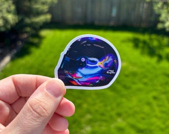 May include: A sticker featuring a colorful abstract design of a racing helmet. The helmet is primarily dark blue with vibrant accents of purple, pink, and yellow. The sticker has a white border and the letters "QL" are visible.