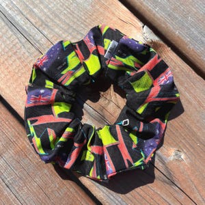 Peut inclure: Un chouchou noir avec un motif vibrant de drapeaux rouges, verts, violets et jaunes. Le chouchou est en tissu et de forme circulaire. Le mot "OSCAR" est visible sur le tissu.