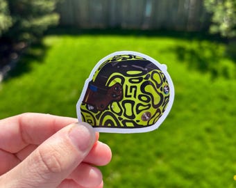 May include: A sticker featuring a racing helmet design. The helmet is black with a yellow, abstract pattern. The visor is brown with a blue strip. The sticker is held in front of a green lawn.