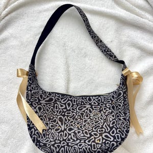 May include: Shoulder bag with a black and white pattern, black strap, and gold ribbon details. The bag has an abstract design and the text "CHAMPION". A fashionable accessory for everyday use.