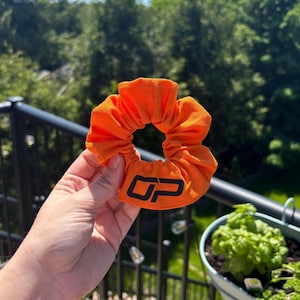 Peut inclure: Chouchou orange vif avec le logo noir "OP". Le tissu est fronc&eacute;, cr&eacute;ant un effet textur&eacute;. Le chouchou est tenu dans une main, avec un arri&egrave;re-plan de feuillage vert.