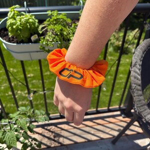 Peut inclure: Chouchou orange vif avec le logo "CP" noir. Le chouchou, en tissu doux, est port&eacute; au poignet. L'arri&egrave;re-plan montre un jardin avec des plantes vertes et une balustrade en m&eacute;tal noir.