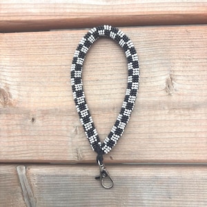 Peut inclure: Une lanière de poignet perlée noire et blanche avec un motif en damier. La lanière a un fermoir de couleur argentée et un cordon noir reliant les perles au fermoir. La lanière est présentée sur une surface en bois.