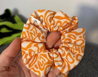 May include: An orange and white patterned scrunchie. The fabric features an abstract design in a repeating pattern. The scrunchie is held in a hand, showcasing its texture and design. A small tag with text is visible.