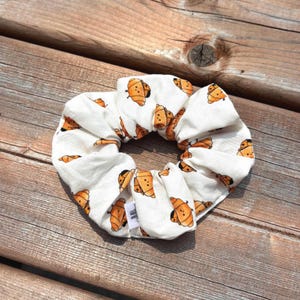 Peut inclure: Chouchou blanc avec un motif répété de croissants orange dessinés. Le tissu est froncé et élastique, créant une forme circulaire. Le chouchou est posé sur une surface en bois.