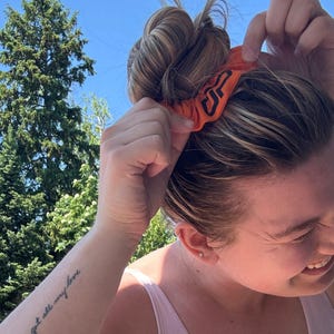 Peut inclure: Un chouchou orange avec un logo noir est plac&eacute; dans un chignon. Le chouchou est fait d'un tissu doux et est de couleur orange vif. Le logo semble &ecirc;tre un design stylis&eacute;.