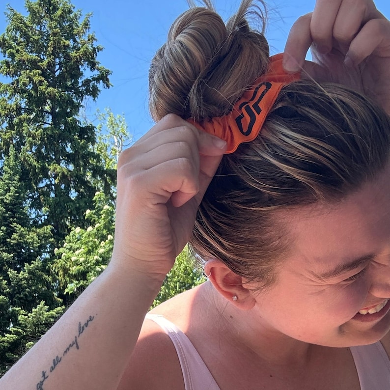 Peut inclure: Un chouchou orange avec un logo noir est plac&eacute; dans un chignon. Le chouchou est fait d'un tissu doux et est de couleur orange vif. Le logo semble &ecirc;tre un design stylis&eacute;.