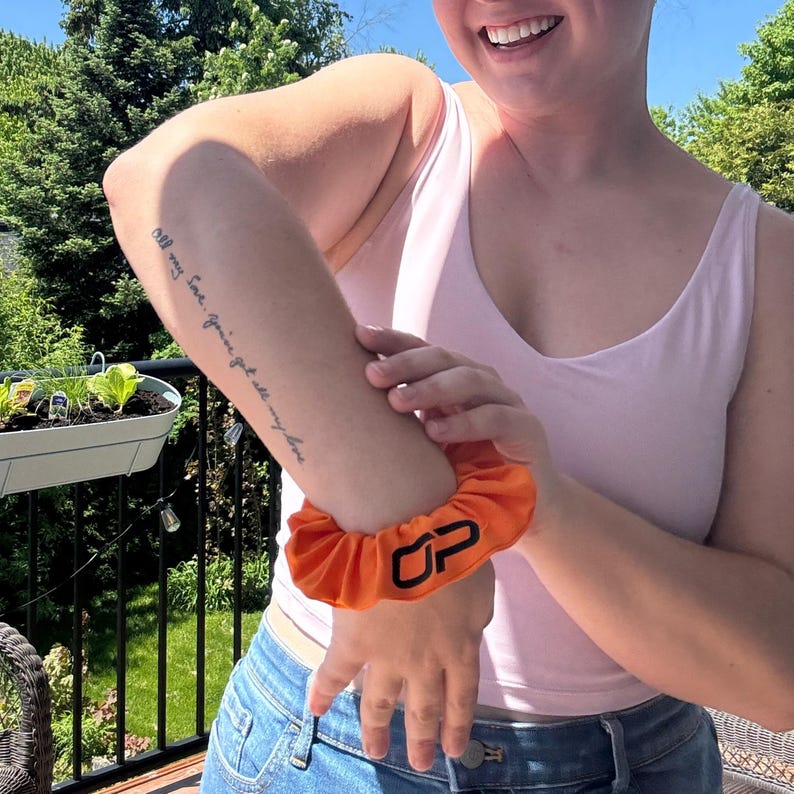 Peut inclure: Un chouchou orange avec les lettres "OP" en noir. Le chouchou est port&eacute; au poignet. La personne porte un d&eacute;bardeur rose et un jean bleu. L'arri&egrave;re-plan comprend un jardin et des arbres.