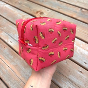May include: A red cosmetic bag featuring a pattern of cartoon hamburgers. It has a red zip and pull tab. The bag is held in a hand.