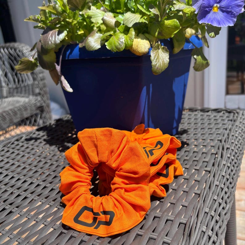 Peut inclure: Des chouchous orange vif avec un logo noir sont pr&eacute;sent&eacute;s sur une surface en osier grise. Un pot de fleurs bleu avec un feuillage vert et une fleur violette est en arri&egrave;re-plan. Les chouchous sont en tissu doux.