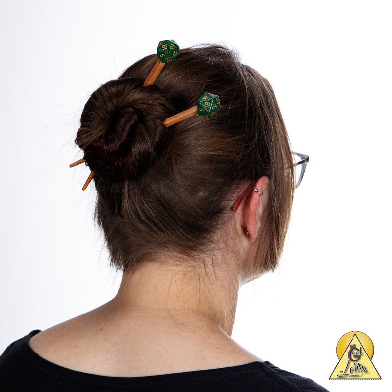 May include: A woman with brown hair in a bun, wearing glasses, with two green and gold dice hair pins in her hair.