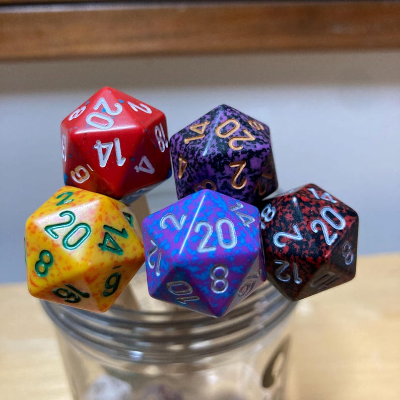 May include: Five 20-sided dice in a glass jar. The dice are red, purple, yellow, blue, and black with white numbers.