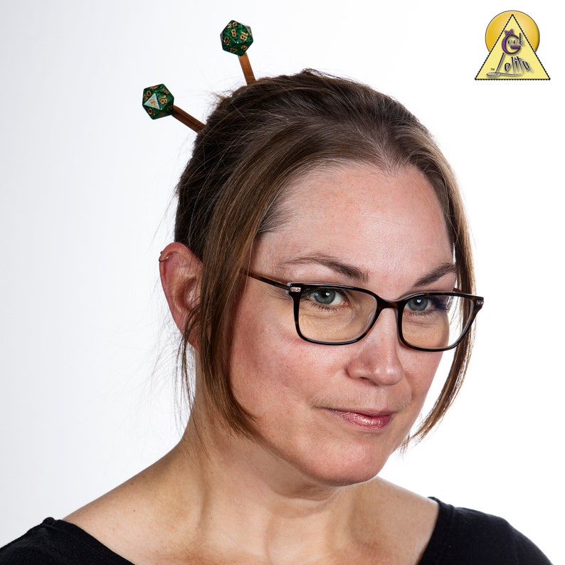 May include: A woman with brown hair is wearing glasses and has two green dice hair pins in her hair.