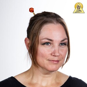 May include: A woman with brown hair wearing a black shirt has a red and black dice on a wooden stick in her hair.
