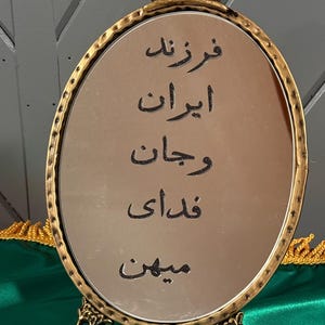 May include: An oval mirror with a gold-colored ornate frame. The mirror has text in a foreign language. The frame has a decorative crown at the top and rests on a decorative base. The background includes green and gold fabric.