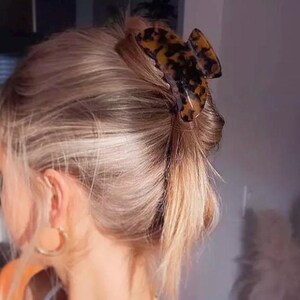 May include: A tortoiseshell hair claw clip holds blonde hair in a casual updo. The clip is a warm brown colour with darker brown spots. Gold hoop earrings are visible.