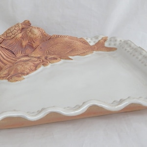 May include: A white ceramic serving dish with a raised design of a turtle, fish, and seaweed. The dish has a scalloped edge and a textured rim.