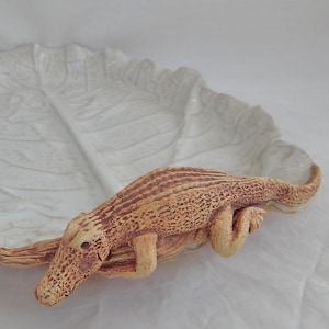 May include: A white ceramic dish shaped like a cabbage leaf with a brown alligator figurine attached to the side.
